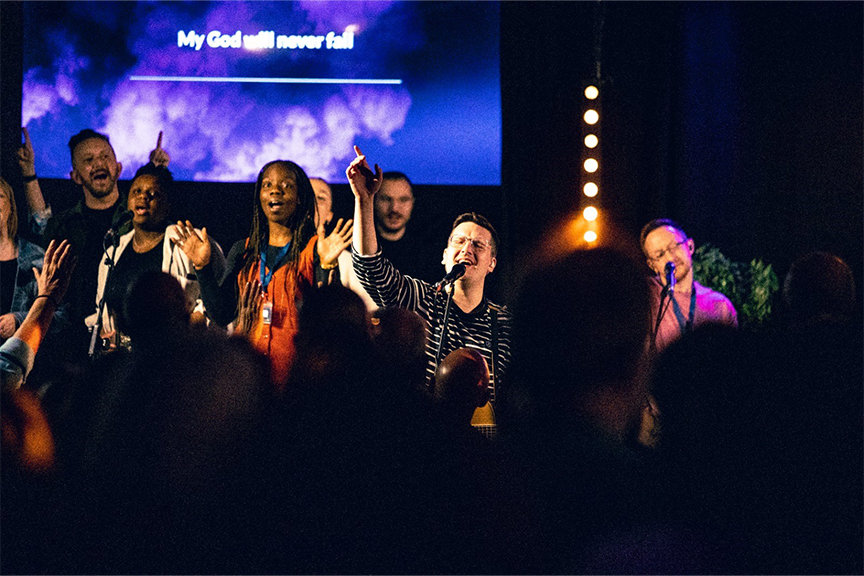 The worship band leading worship in a dark, ambient room with lights and a colourful screen behind them