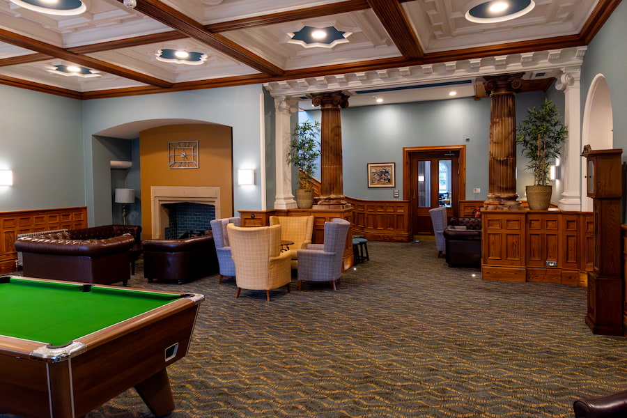 The Hayes Lounge today with a pool table, the same fireplace, comfy chairs, and the same marble pillars.