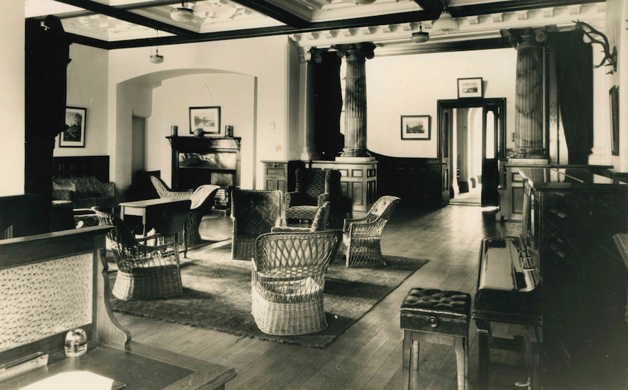 The Hayes Lounge in 1920 with old wicca chairs, a fireplace, and big marble pillars.